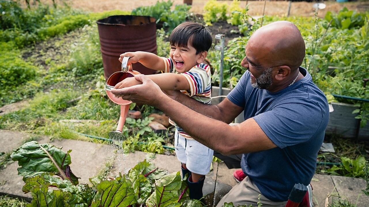 Man helping a smiling child water plants in a garden