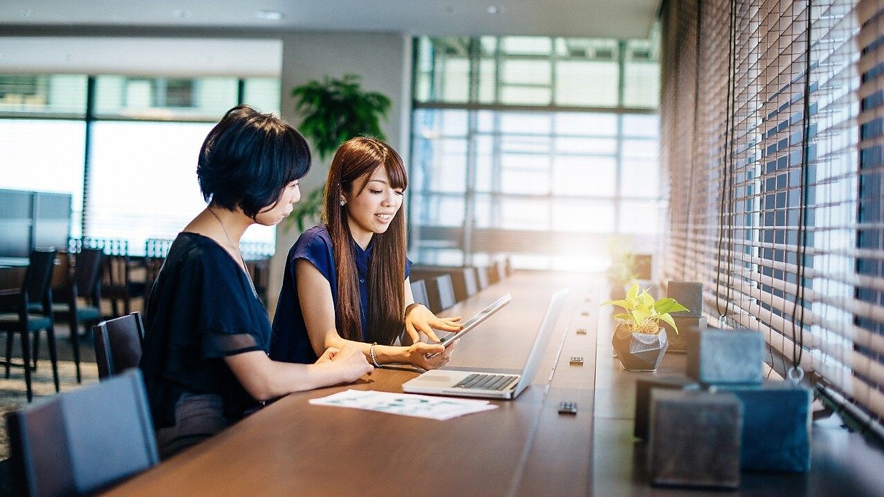 Two women sitting at a desk looking at a computer and a tablet