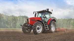 A red tractor ploughing a muddy field with trees in the background
