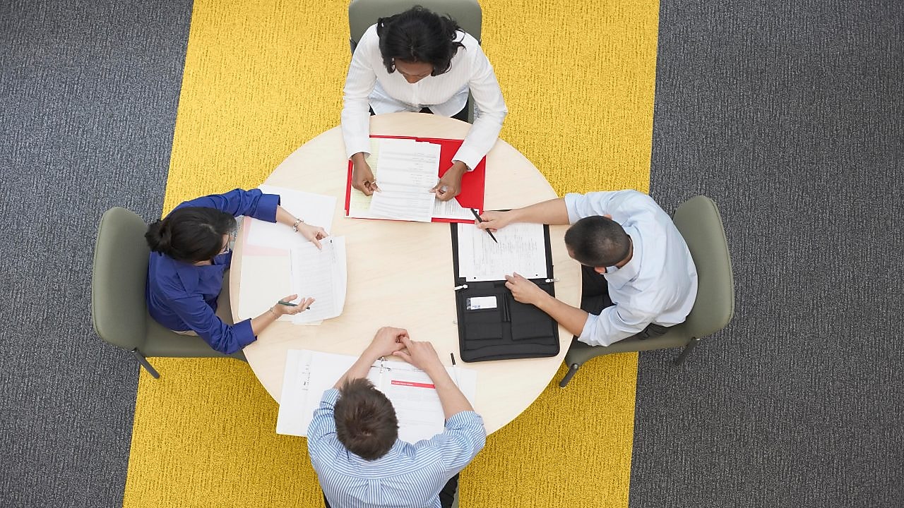 Four business people sitting around a round table reading documents, grey and yellow carpet