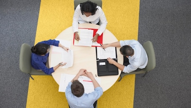 Four business people sitting around a round table reading documents, grey and yellow carpet