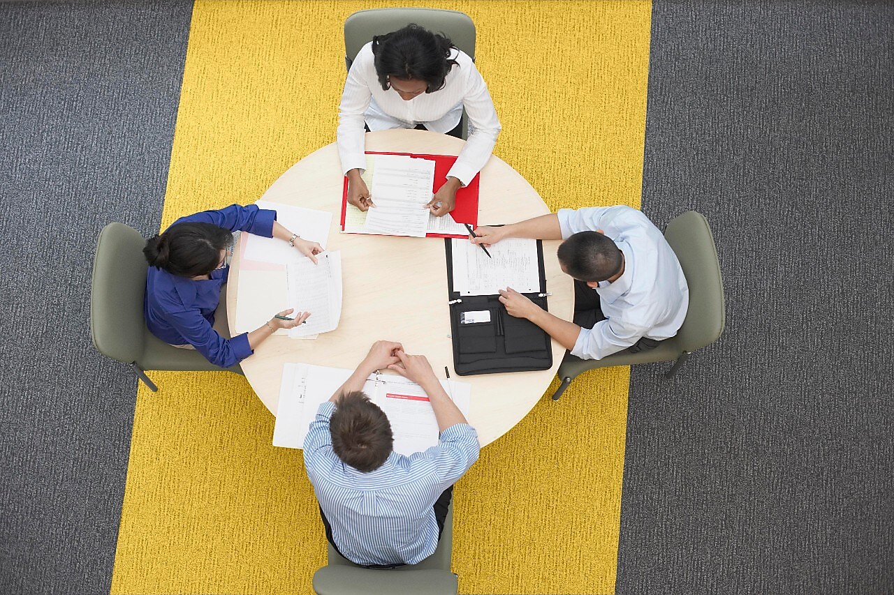 Four business people sitting around a round table reading documents, grey and yellow carpet